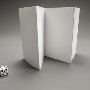 Puede incluir: Una pantalla de papel blanco plegada con un solo dado blanco con puntos negros en primer plano.