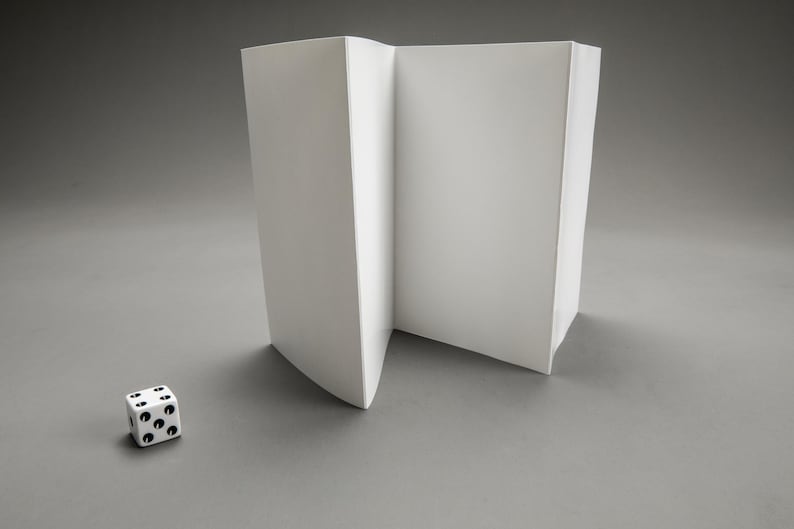 Puede incluir: Una pantalla de papel blanco plegada con un solo dado blanco con puntos negros en primer plano.