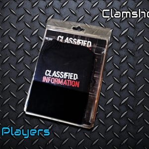 May include: A black product in a clear clamshell package with the words "CLASSIFIED INFORMATION" in white and red. The package is on a textured, dark metal surface. The text "2 Players" is in blue.