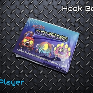 May include: A boxed game titled "Micro Disturblings" with cartoon monster illustrations. The box is wrapped in plastic and has the text "Hook Box!" and "1 Player". The background is a textured, dark metal surface.