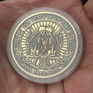 Könnte beinhalten: Eine goldfarbene Münze mit einem Design von zwei Skeletten auf einer Schaukel, umgeben von Rosen. Die Münze befindet sich in einer transparenten Kunststoffhülle. Der Text auf der Münze lautet "TILL DEATH DO US PART" und "AND THEN SOME".