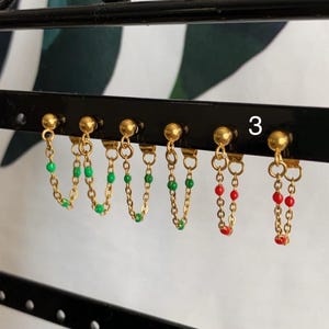 Op de afbeelding: Goudkleurige oorbellen met een kettingontwerp. De oorbellen hebben kleine, kleurrijke kralen in groen en rood. De oorbellen worden weergegeven op een zwart rek met het nummer "3" zichtbaar.