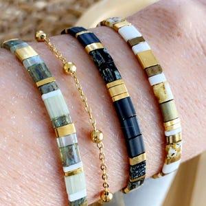 Peut inclure: Trois bracelets de perles avec des accents dorés sont présentés sur un poignet. Les bracelets présentent des perles blanches, noires et vertes, séparées par des entretoises en métal doré. Une délicate chaîne dorée avec de petites perles relie deux des bracelets.