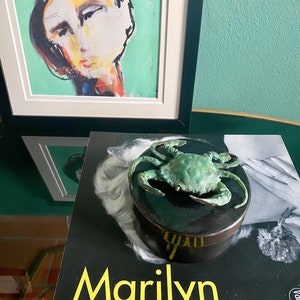 May include: A black and white book with the title "Marilyn" in yellow letters. A green ceramic box with a bronze crab sculpture on top is on the book.