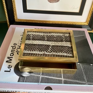 以下が含まれることがあります： 蓋にヘビの皮の模様が施された金色調の箱。箱には引き出しがあり、開いています。箱は、「Le Monde d'Henri Cartier-Bresson」という文字が表紙に印刷された本の上に置かれています。