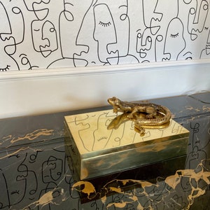 Peut inclure: Une boîte en métal doré avec une figurine de lézard sur le dessus. La boîte est posée sur une surface en marbre noir et or. L'arrière-plan est un mur blanc avec un motif abstrait noir et blanc.