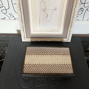 以下が含まれることがあります： 右下に署名のある、顔の白黒線画が額装されています。額縁は白で、装飾的なディテールが施されています。蛇の皮模様の黒い表面の上に置かれています。