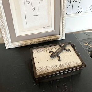 以下が含まれることがあります： 黒いトカゲが乗った小さな白い箱。箱は蛇の皮の模様で、質感があります。箱は黒い表面の上に置かれています。