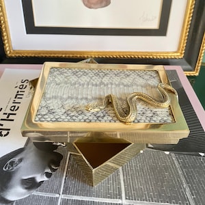 May include: A gold-coloured box with a snake design on the lid. The box is made of metal and has a snakeskin pattern on the lid. The box is open and the inside is visible. The box is sitting on a magazine.