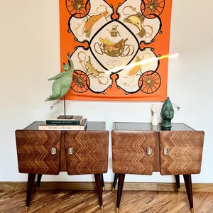 以下が含まれることがあります： ダークウッドのヘリンボーン柄のドア、黒い天板、真鍮の先端が付いた脚を備えた、ミッドセンチュリーモダン様式のナイトスタンド2つ。緑色の彫刻と、馬車のデザインが施された装飾的なスカーフが上に吊り下げられています。