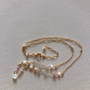 Puede incluir: Un collar de cadena dorada con una variedad de cuentas de colores pastel, incluyendo rosa, azul y blanco. El collar est&aacute; superpuesto con una segunda cadena dorada.