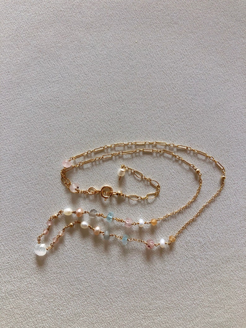 Puede incluir: Un collar de cadena dorada con una variedad de cuentas de colores pastel, incluyendo rosa, azul y blanco. El collar est&aacute; superpuesto con una segunda cadena dorada.