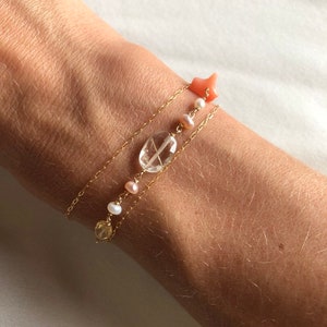 May include: Three gold chain bracelets with different charms. The top bracelet has a pink coral star charm. The middle bracelet has a clear quartz oval charm and pink pearl beads. The bottom bracelet has a yellow citrine charm and white pearl beads.