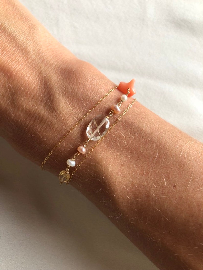 May include: Three gold chain bracelets with different charms. The top bracelet has a pink coral star charm. The middle bracelet has a clear quartz oval charm and pink pearl beads. The bottom bracelet has a yellow citrine charm and white pearl beads.