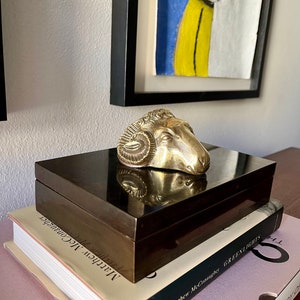 Könnte beinhalten: Eine Skulptur eines Widderkopfes aus Messing steht auf einer dunkelbraunen Holzkiste. Die Kiste steht auf zwei Büchern, eines mit dem Titel "Greenlights" von Matthew McConaughey.