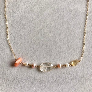 May include: A gold chain necklace with a variety of gemstone beads, including a clear oval-shaped stone, two small white pearls, a small pink stone, and a small yellow stone.