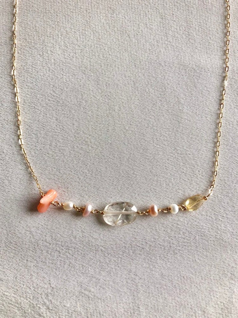 May include: A gold chain necklace with a variety of gemstone beads, including a clear oval-shaped stone, two small white pearls, a small pink stone, and a small yellow stone.