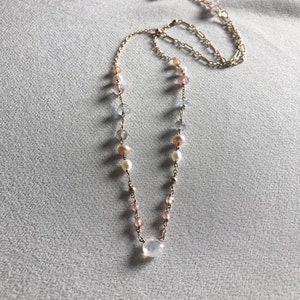 Puede incluir: Un delicado collar de cadena dorada con una variedad de cuentas de colores pastel, incluyendo rosa, azul y blanco. El collar presenta un colgante de piedra preciosa transparente m&aacute;s grande.