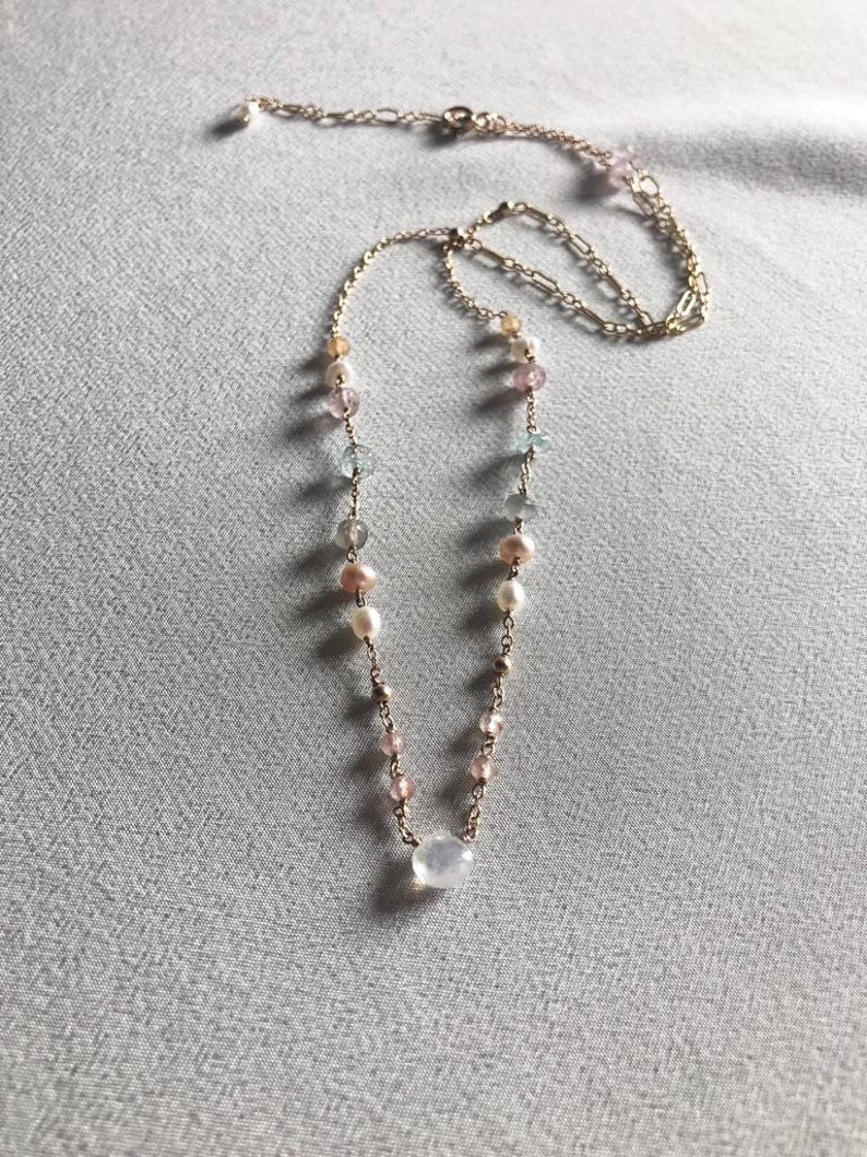 Puede incluir: Un delicado collar de cadena dorada con una variedad de cuentas de colores pastel, incluyendo rosa, azul y blanco. El collar presenta un colgante de piedra preciosa transparente m&aacute;s grande.