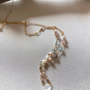 Puede incluir: Un collar de cadena dorada con una variedad de cuentas de piedras preciosas, incluyendo perlas, piedras rosas, azules y grises. El collar tiene un colgante de piedra de luna en forma de l&aacute;grima.