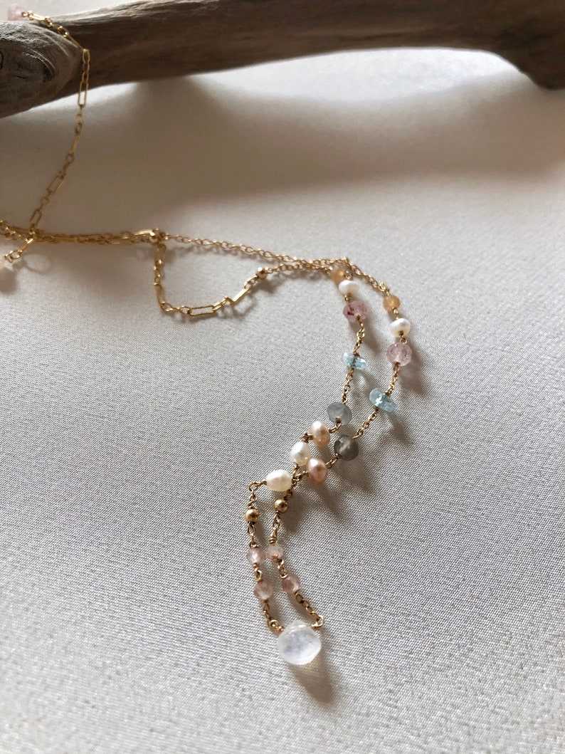 Puede incluir: Un collar de cadena dorada con una variedad de cuentas de piedras preciosas, incluyendo perlas, piedras rosas, azules y grises. El collar tiene un colgante de piedra de luna en forma de l&aacute;grima.