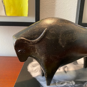 Puede incluir: Una escultura estilizada de toro marr&oacute;n oscuro con un acabado texturizado. El toro est&aacute; de pie sobre una superficie negra con la palabra "Marilyn" visible en el fondo.