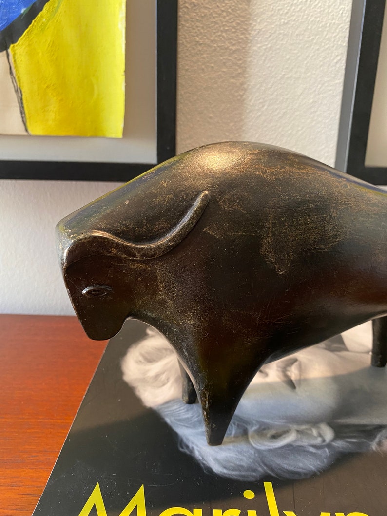 Puede incluir: Una escultura estilizada de toro marr&oacute;n oscuro con un acabado texturizado. El toro est&aacute; de pie sobre una superficie negra con la palabra "Marilyn" visible en el fondo.