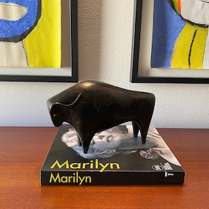 Puede incluir: Una escultura de bronce negra en forma de toro sobre un libro negro con el t&iacute;tulo "Marilyn Marilyn" en letras doradas.