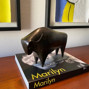 Puede incluir: Una escultura de bronce negro de un toro sobre un libro negro con el t&iacute;tulo "Marilyn" en letras amarillas.