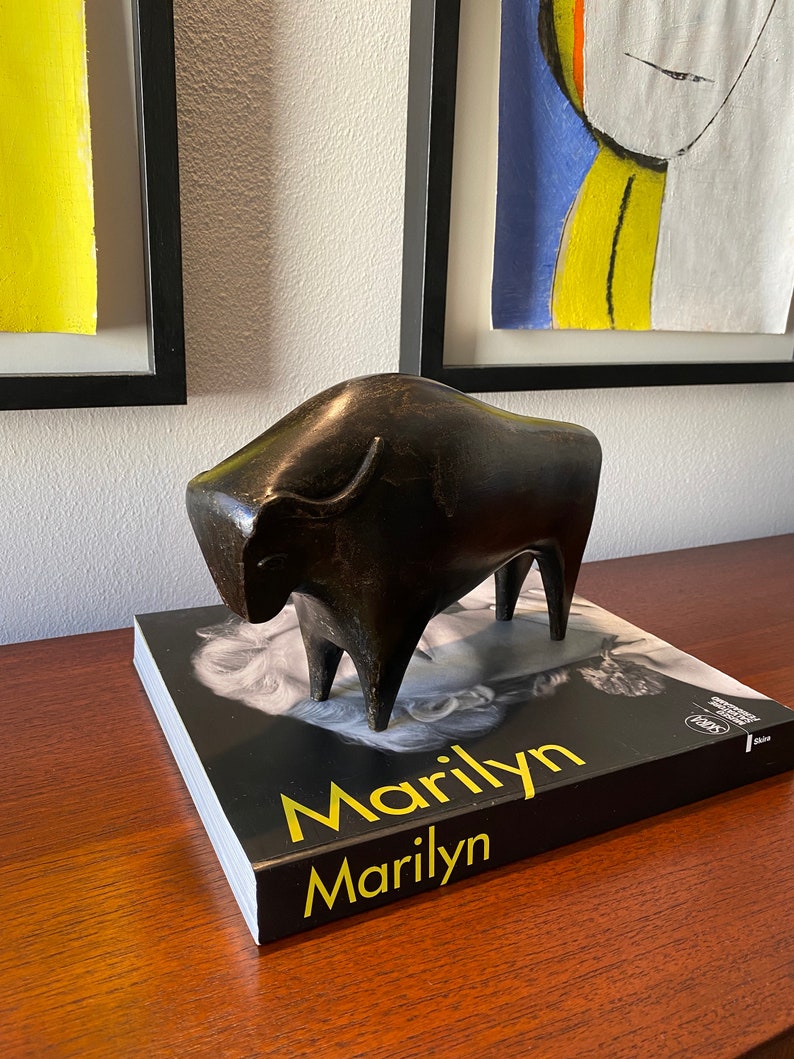 Puede incluir: Una escultura de bronce negro de un toro sobre un libro negro con el t&iacute;tulo "Marilyn" en letras amarillas.