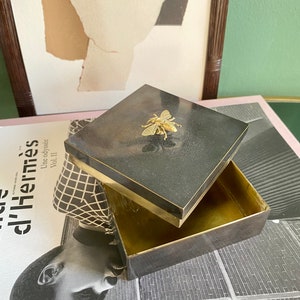 May include: A black square box with a gold trim and a gold bee design on the lid. The box is open and the inside is empty.