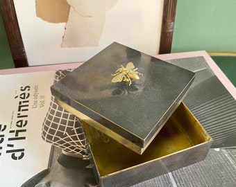 May include: A black square box with a gold trim and a gold bee design on the lid. The box is open and the inside is empty.