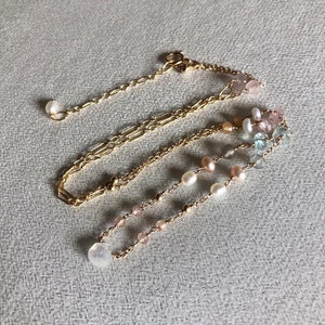 Puede incluir: Un collar de cadena dorada con m&uacute;ltiples hilos de cuentas de diferentes colores, incluyendo rosa, blanco y azul. El collar est&aacute; sobre un fondo de tela gris.