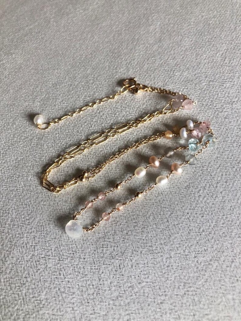 Puede incluir: Un collar de cadena dorada con m&uacute;ltiples hilos de cuentas de diferentes colores, incluyendo rosa, blanco y azul. El collar est&aacute; sobre un fondo de tela gris.