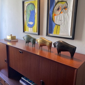 Puede incluir: Un aparador de madera con tres cajones y dos puertas. El aparador est&aacute; decorado con cuatro figuritas de animales de bronce y una peque&ntilde;a caja dorada en la parte superior. Dos pinturas abstractas enmarcadas de rostros est&aacute;n en la pared detr&aacute;s del aparador.