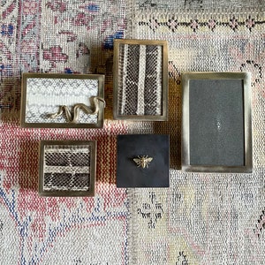 Peut inclure: Quatre petites boîtes décoratives avec des motifs différents. Les boîtes sont en bois et ont une finition en laiton. Une boîte a un motif de serpent, une autre a un motif d'abeille, une autre a un motif de peau de serpent et une autre a un motif texturé.