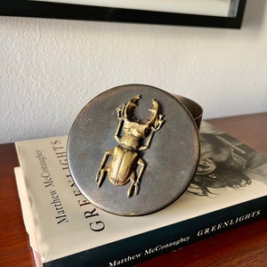 Puede incluir: Una caja redonda de color plateado con un diseño de escarabajo dorado en la tapa. La caja está encima de un libro titulado "Greenlights" de Matthew McConaughey.