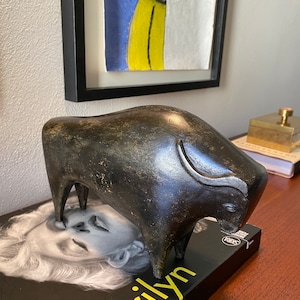 Puede incluir: Una escultura estilizada de un toro gris oscuro, de pie sobre un libro con la palabra "Marilyn" impresa en la portada.