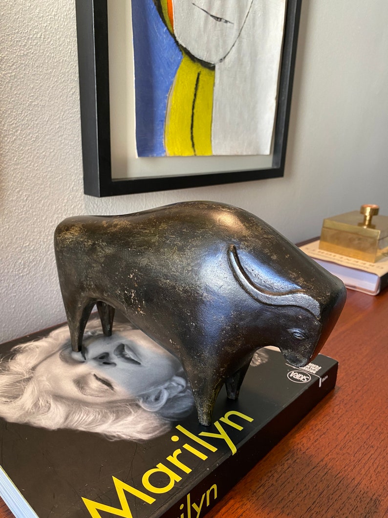 Puede incluir: Una escultura estilizada de un toro gris oscuro, de pie sobre un libro con la palabra "Marilyn" impresa en la portada.