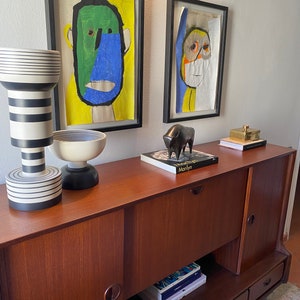 Puede incluir: Un armario de madera con dos puertas y tres cajones. El armario est&aacute; decorado con dos cuadros enmarcados de caras abstractas y una escultura de toro negro. El armario est&aacute; sobre una alfombra con un patr&oacute;n geom&eacute;trico.