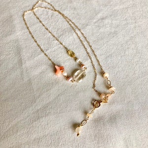 May include: A delicate gold chain necklace with a pink and white gemstone pendant. The pendant features a clear crystal, a pink star, and small pink and white beads.