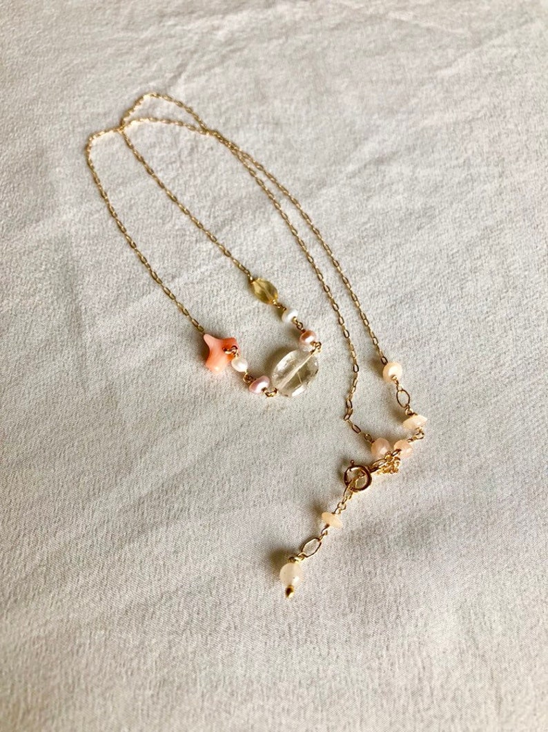 May include: A delicate gold chain necklace with a pink and white gemstone pendant. The pendant features a clear crystal, a pink star, and small pink and white beads.