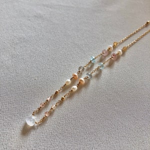 Puede incluir: Un collar de cadena dorada con una variedad de cuentas de piedras preciosas, incluyendo rosa, azul y blanco. El collar presenta un gran colgante de piedra lunar blanca.