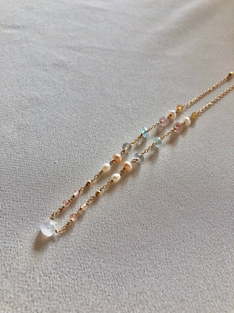 Puede incluir: Un collar de cadena dorada con una variedad de cuentas de piedras preciosas, incluyendo rosa, azul y blanco. El collar presenta un gran colgante de piedra lunar blanca.