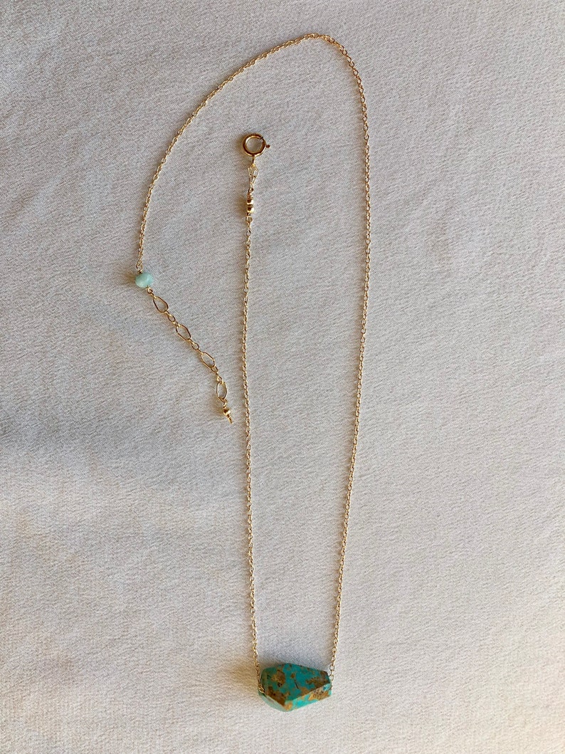 May include: A gold chain necklace with a turquoise stone pendant. The stone is a rough, irregular shape and has a green and brown colour.
