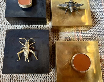 May include: Four decorative boxes with different designs. Two boxes are black with brass accents, one has a leather patch and the other has a brass cricket. Two boxes are gold with brass accents, one has a leather patch and the other has a brass bee.