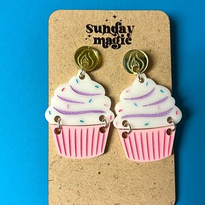Puede incluir: Un par de pendientes con forma de cupcake con glaseado rosa y blanco, chispas y detalles dorados. Los pendientes est&aacute;n en una tarjeta marr&oacute;n con el texto "Sunday Magic".