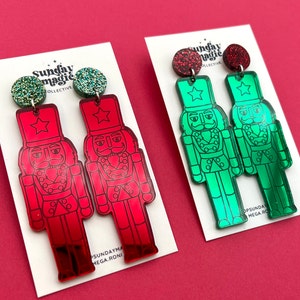 Może przedstawiać: Para czerwonych i zielonych lustrzanych kolczyków w kształcie postaci z łamacza orzechów z błyszczącymi akcentami. Kolczyki znajdują się na białej kartce z tekstem "Sunday Magic Collective".