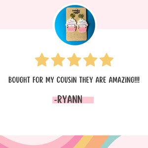 Puede incluir: Cinco estrellas doradas y el texto "BOUGHT FOR MY COUSIN THEY ARE AMAZING!!!" con el nombre "RYANN" debajo. La imagen tambi&eacute;n muestra un par de pendientes de cupcake rosa y blanco.