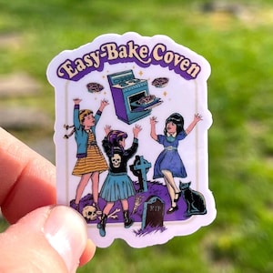 May include: A sticker with the text "Easy-Bake Coven" features three girls celebrating around a purple oven. The illustration includes a black cat, a tombstone, and a skull, with a green background.